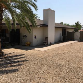 A single-story house with a chimney and a driveway, surrounded by a desert landscape with a palm tree.