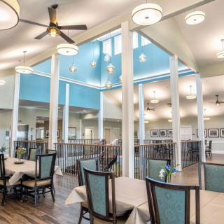 A spacious and bright dining area with modern decor, featuring tables and chairs arranged neatly under a high ceiling with fans.
