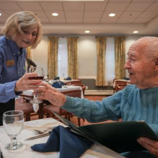 An elderly man enjoying a meal as a waitress serves him wine in a restaurant setting.