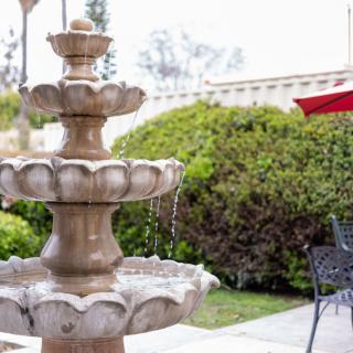 A garden scene featuring a multi-tiered fountain with flowing water, surrounded by lush greenery and patio furniture.