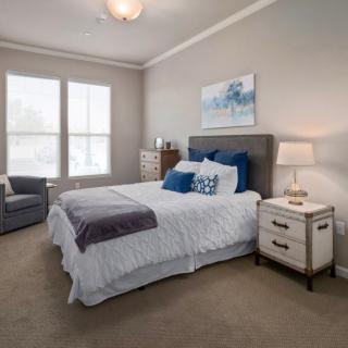 A cozy bedroom featuring a neatly made bed with blue and white bedding, flanked by a nightstand with a lamp. A comfortable chair is placed by the window, and artwork adorns the wall.