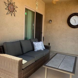 A cozy patio setup featuring a wicker sofa with cushions, a coffee table, and wall decorations including a clock and sun ornament.