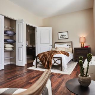 A cozy bedroom with hardwood floors, featuring a neatly made bed, a nightstand with a lamp, and an open closet.