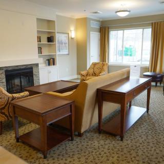A cozy living room with a sofa, chairs, and a fireplace, featuring a bookshelf and large window.
