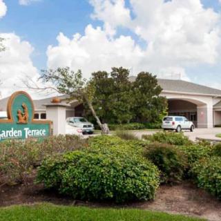 An exterior view of a building entrance with landscaped greenery and parked vehicles under a partly cloudy sky.