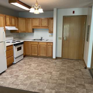 An empty kitchen area with wooden cabinets and tile flooring, featuring a refrigerator, oven, and sink.