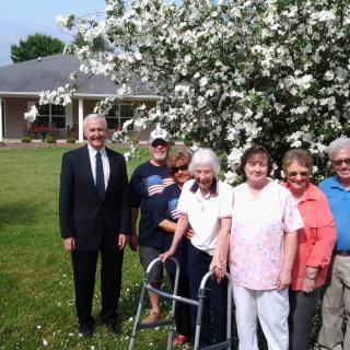 A group of people, including elderly individuals, pose together outdoors near a blooming tree.