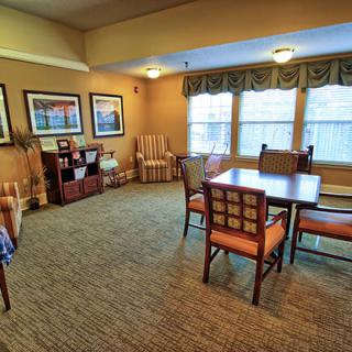 A cozy living room with a table and chairs, carpeted floor, artwork on the walls, and large windows.