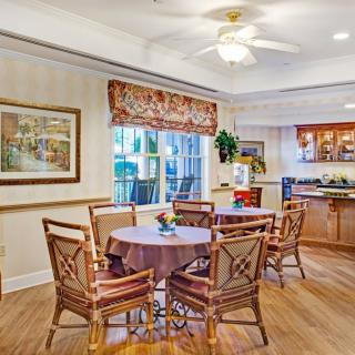 A cozy dining area with a round table and wooden chairs, featuring a nearby kitchen and a window with decorative blinds.