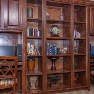 A home office with a wooden bookshelf filled with books, decorative items, and a clock alongside a computer desk.