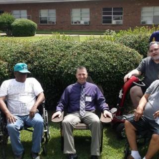 A group of people, some in wheelchairs, sitting outdoors near bushes and a building.