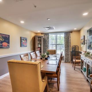 A cozy dining room featuring a long wooden table with chairs, decorated with artwork on the walls and large windows adorned with patterned curtains.