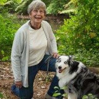 A woman kneeling in a forested area next to a dog, both appearing happy and relaxed.