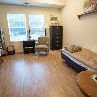 A cozy room with wooden flooring featuring a bed, chairs, a cabinet, and a window allowing natural light.