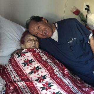 An elderly person lying in bed, wrapped in a festive blanket, accompanied by a smiling individual showing affection.