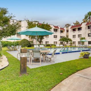 A hotel courtyard with a swimming pool, surrounded by tables and chairs with umbrellas, amidst well-maintained greenery.