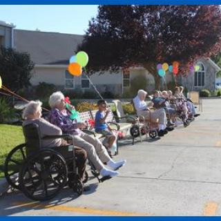A group of elderly people enjoying a sunny day outside, sitting on benches and in wheelchairs, with colorful balloons around them.