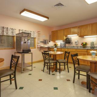 A cozy dining area with wooden tables and chairs, featuring a kitchen setup with cabinets and overhead lighting.