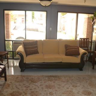 A cozy living room featuring a beige sofa, wooden chairs, and ample natural light.