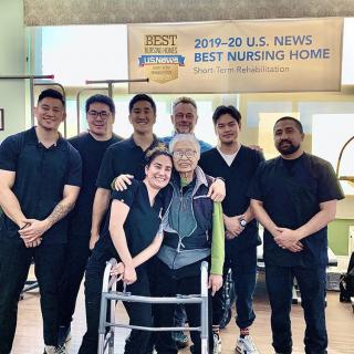 A group of healthcare staff posing with an elderly person, standing in front of a 'Best Nursing Home' award banner.