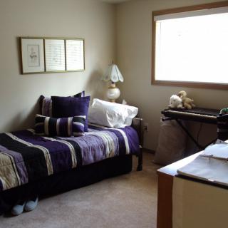 A cozy bedroom with a single bed featuring striped bedding, decorative pillows, and a lamp by the window. A desk is positioned in front of the window.