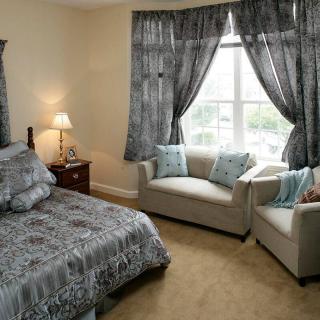 A cozy bedroom featuring a bed with patterned bedding, armchairs by the window with matching curtains, and a bedside lamp providing warm light.