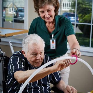 An elderly man participates in a therapy session with assistance from a caregiver.
