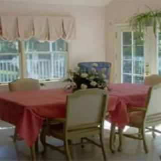 A bright dining room with a table set for four, featuring pink tablecloths and a floral centerpiece near large windows.