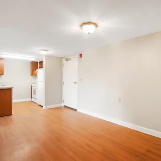 A view of an empty apartment interior featuring a kitchen area and living space with wood flooring and white walls.