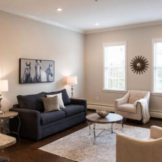 A modern living room with neutral tones, featuring a dark gray sofa, two armchairs, and a central coffee table. The space is accentuated by wall art and a sunburst mirror.