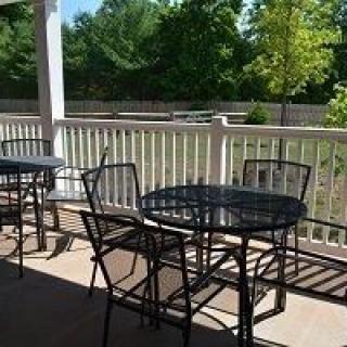 A sunny patio area with metal tables and chairs, surrounded by a white fence and green trees in the background.