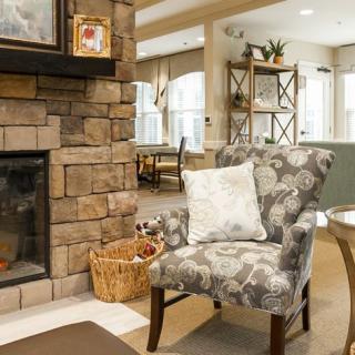 A cozy living room featuring a stone fireplace, an upholstered chair with a cushion, and a warm, inviting atmosphere.
