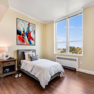 A modern bedroom with a single bed and colorful artwork above, featuring large windows and hardwood flooring.