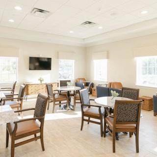 A bright and airy dining area with multiple tables and chairs, featuring a wall-mounted television and large windows.