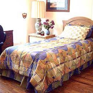 A cozy bedroom featuring a single bed with colorful bedding, a nightstand with a lamp and a vase of flowers.
