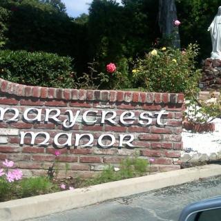 A brick sign for 'Marycrest Manor' surrounded by flowers and a statue.