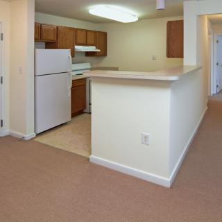 A modern apartment interior featuring a kitchen with wooden cabinets and a white fridge, adjacent to a hallway.