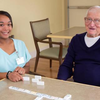A nurse and a senior man enjoying a game of dominoes together at a table.