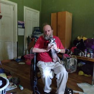 A person in a wheelchair holding a small dog in a cluttered indoor space.