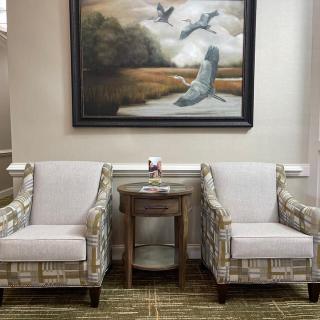 A cozy living room setup with two patterned armchairs flanking a small round table, beneath a large framed painting of birds over a landscape.