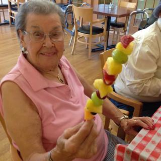 An elderly woman smiling while holding a colorful fruit skewer at a restaurant.