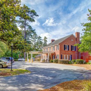 A brick building surrounded by trees with a parking lot in front, under a bright blue sky.