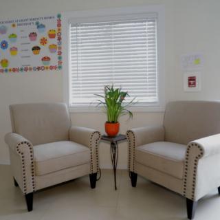 A cozy waiting area with two beige armchairs and a small table with a potted plant, set against a window with blinds.