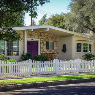 A quaint single-story house with a white picket fence and surrounded by trees.