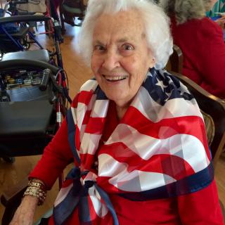 An elderly woman wearing a scarf with the American flag pattern and a red outfit, smiling warmly.