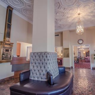 A classic hotel lobby featuring a decorative ceiling and chandelier, with comfortable seating around a central pillar.