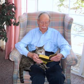 An elderly man sitting in an armchair, reading a book with a cat on his lap.