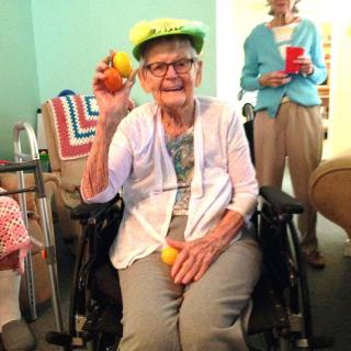 An elderly woman in a wheelchair smiling and holding colorful balls, wearing a bright hat during a celebration.