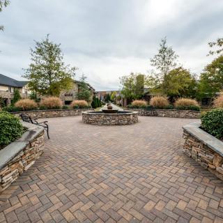 A peaceful courtyard with a central fountain, surrounded by trees and benches on a brick-paved area.