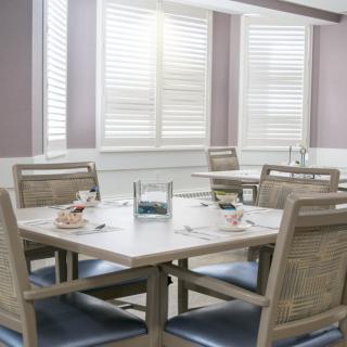 A modern dining room with a white table and cushioned chairs, complemented by window shutters.
